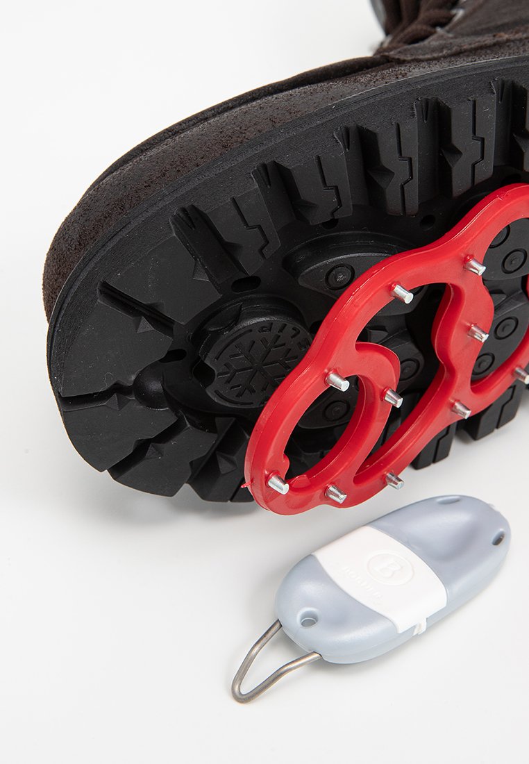 Schwarze Gummistiefelsohle mit auffälligem Profilmuster, rohes Zuggerät angebracht und ein graues sowie weißes Clip-Zubehör daneben.