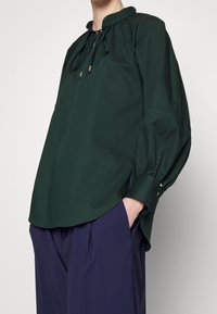 Personne portant un chemisier vert foncé à manches longues avec un col à cordon et un pantalon large bleu marine, main dans la poche, debout sur fond blanc.