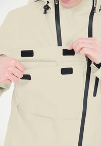 Beige wasserdichte Jacke mit zwei mit Klettverschluss gesicherten Fronttaschen und schwarzen Reißverschlüssen. Glatte Textur mit verstellbarem Kapuze und Kordelzügen.