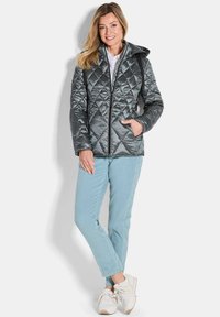 Gepolsterte graue Pufferjacke mit Kapuze, die diagonale Nähte und zwei seitliche Taschen hat, kombiniert mit hellblauen Hosen und weißen Turnschuhen.