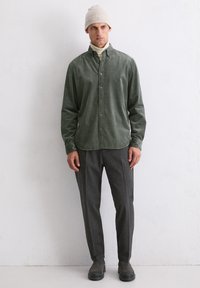Grüne Cord-Hemdjacke, graue strukturierte Hose, schwarze Knöchelstiefel und eine leichte Strickmütze. Schlichter Hintergrund.