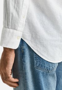Weißes Leinenhemd mit knöpfbarem Manschette, kombiniert mit blauen Jeans. Das Hemd hat eine weiche Textur und einen geschwungenen Saum.