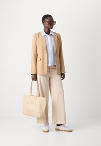 Blazer beige sur chemise bleu clair, pantalons larges crème, baskets blanches avec des accents beige, et un sac fourre-tout à motifs beige clair.
