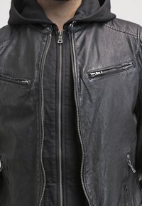Schwarze Lederjacke mit Reißverschluss vorne, zwei Reißverschlusstaschen auf der Brust und einer strukturierten Oberfläche, über einem schwarzen Hoodie getragen.