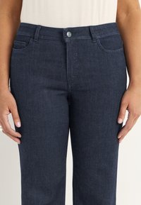 Närmre bild av en person som bär mörkblå denimjeans med knapp och dragkedja, med båda händerna vilande vid sidorna.