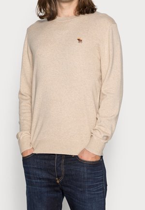 Personne portant un pull beige avec un petit logo d'élan sur la poitrine et un jean bleu foncé, les mains dans les poches, debout devant un fond uni.