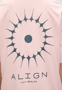 Ροζ πουκάμισο με μαύρο αφηρημένο κυκλικό σχέδιο ηλιακής ακτινοβολίας και τη λέξη «ALIGN» πάνω από το «LALA BERLIN» τυπωμένα στο πίσω μέρος.