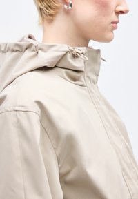 Άτομο με κοντά ξανθά μαλλιά που φοράει μπεζ μπουφάν με κουκούλα και ρυθμιζόμενο κορδόνι, καθώς και ασημένιο σκουλαρίκι κρίκο, βλέπει προς τα δεξιά.