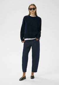 Námornícky pletený sveter, uvoľnený strih, dlhé rukávy; kombinovaný s tmavými džínsami a čiernymi mokasínami. Modelka má na sebe tmavé slnečné okuliare.