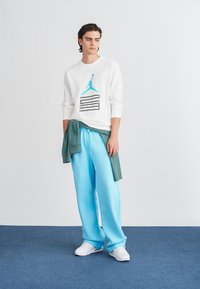 Baltā sporta krekla ar zilo Jordan grafiku, kombinācijā ar gaiši ziliem treniņbiksēm un zaļu džemperi, kas sasietas ap vidu. Baltas sporta kurpes.