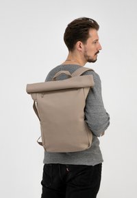 Rucsac bej cu capac tip rulou, fabricat din material rezistent, dotat cu un buzunar cu fermoar în față și bretele ajustabile, purtat pe umăr.