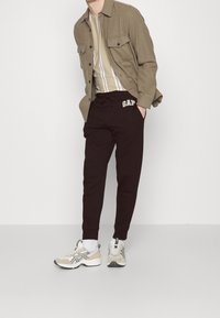 Braune Shirtjacke über einem beige-weißen gestreiften T-Shirt, kombiniert mit dunkelburgunderfarbenen Sweatpants mit "GAP"-Logo und beigen Sneakern.