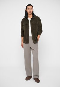 Olijfgroene corduroy overshirt met twee borstzakken, gecombineerd met lichtgrijze geplooide broek en bruine veterschoenen. Kenmerkt zich door gladde texturen.