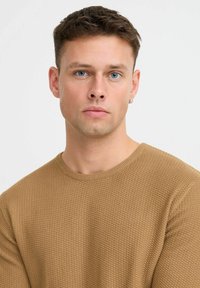 Suéter marrón texturizado con un patrón de canalé, escote redondo y un diseño ajustado. El modelo tiene el cabello corto y lleva un arete.