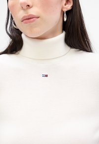 Pulover cu guler înalt de culoare crem, având o textură moale și un mic logo brodat în nuanțe de navy, roșu și alb pe piept.