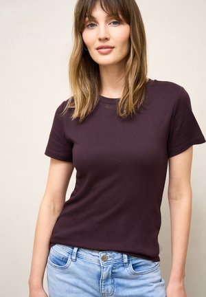 Kvinde med skulderlangt, lysebrunt hår, iført en mørkelilla t-shirt med korte ærmer og lyseblå jeans mod en ensfarvet baggrund.