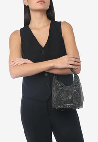 Zwarte handtas gemaakt van sprankelende rhinestones, met een gebogen vorm en kleine handvatten, vastgehouden in de arm van een model met een op maat gemaakt zwart vest.