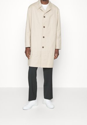 Beige cotton trench coat with a collar, front buttons, and side pockets, paired with dark trousers and white sneakers.