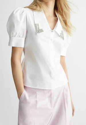 Witte blouse met korte mouwen en pofmouwen, knoopsluiting aan de voorkant en een versierde kraag met kristallen accenten. Gecombineerd met lichtroze broeken.