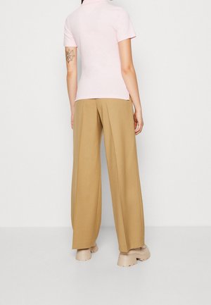 Person trägt ein hellrosa kurzärmeliges Oberteil und weite beige Hosen, steht barfuß an einer schlichten weißen Wand.