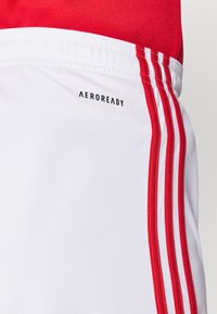 Fehér sportnadrág piros oldalsó csíkokkal és "AEROREADY" logóval. Sima anyag modern szabással. További részletek nem láthatók.