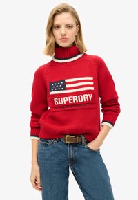 Rode gebreide trui met hoge hals, voorzien van een ontwerp van de Amerikaanse vlag en "SUPERDRY" merknaam. Ribgebreide manchetten met blauwe en crèmekleurige accenten.