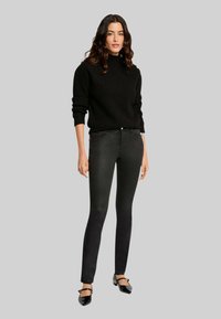Suéter negro de punto con un alto cuello y mangas largas, combinado con jeans negros ajustados. La modelo lleva bailarinas negras. Fondo neutro.