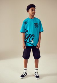 T-shirt turquoise oversize avec texte et accents noirs de style graffiti, assorti à un short noir et des baskets noires à semelles blanches avec des chaussettes.