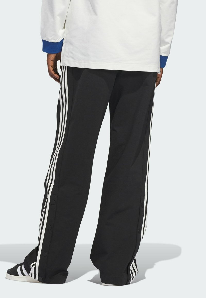 Black track pants with white stripes along the sides, featuring snap-button closures and a loose fit. Textured fabric, paired with a white top.