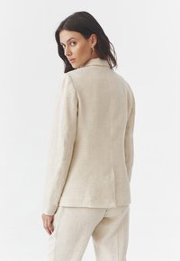 Beige len blézer, szabott fazonú, hosszú ujjú, felső gallérral és hátsó szellőzőnyílással, hozzáillő nadrággal párosítva. Sima textúra.