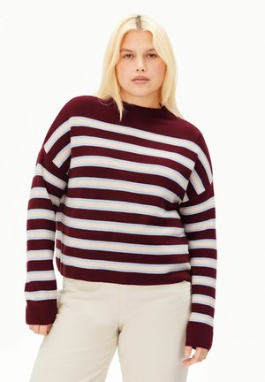 Jeune personne aux longs cheveux blonds portant un pull à manches longues rayé bordeaux et blanc et un pantalon clair sur fond blanc.