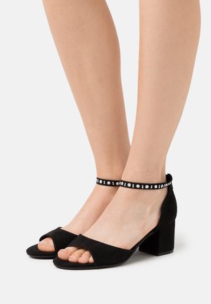 Sandales à talons en daim noir avec design à bout ouvert, talon bloc épais et bride de cheville ornée de petits accents circulaires.