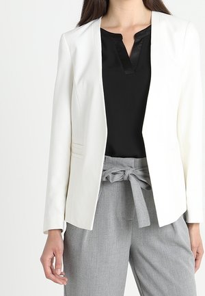 Vrouw die een witte blazer draagt, een zwarte blouse met een V-hals en een grijze broek met hoge taille, vastgebonden met een stoffen strikriem in de taille.