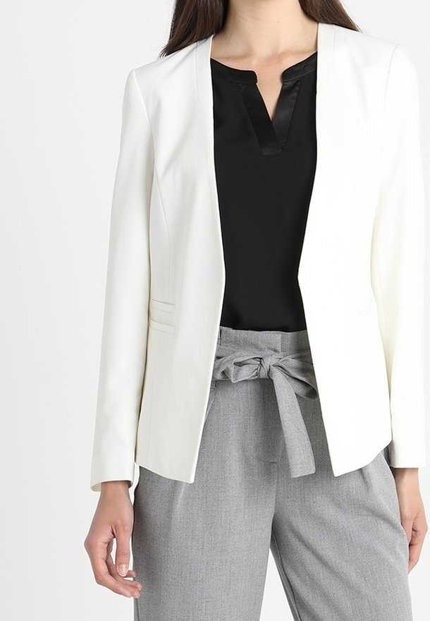 Femme portant un blazer blanc, un chemisier noir à col en V et un pantalon gris taille haute noué à la taille avec un nœud en tissu.