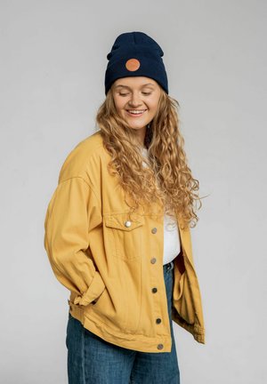 Żółta dżinsowa kurtka z srebnymi guzikami, noszona na białej koszuli. Granatowy beanie z pomarańczową łatką dopełnia strój. Widoczne długie, kręcone włosy.