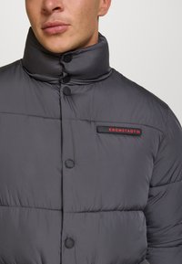 Primo piano di una persona che indossa un giaccone grigio imbottito con colletto alto, bottoni neri e un logo rosso "KRONSTADT" sul petto.