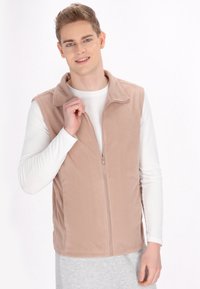 Gilet in pile beige chiaro con zip frontale, collo a camicia e texture liscia. Indossato sopra una maglietta bianca a maniche lunghe.