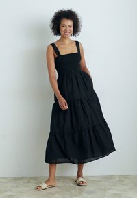 Svart, ermeløs maxi-kjole med firkantet halslinning, lagdelt design og smocket bodice. Modellen har på seg lette sandaler på et mønstret gulv.
