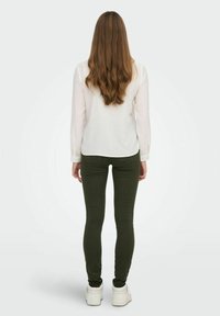 Chemise blanche à manches longues et jean skinny vert foncé avec une texture lisse ; modèle de dos, portant des baskets blanches, avec de longs cheveux ondulés.