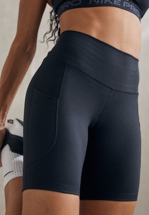 Frau trägt schwarze, hoch taillierte Sportshorts mit Seitentasche und schwarzen Nike Pro Sport-BH und dehnt das Bein hinter dem Körper.