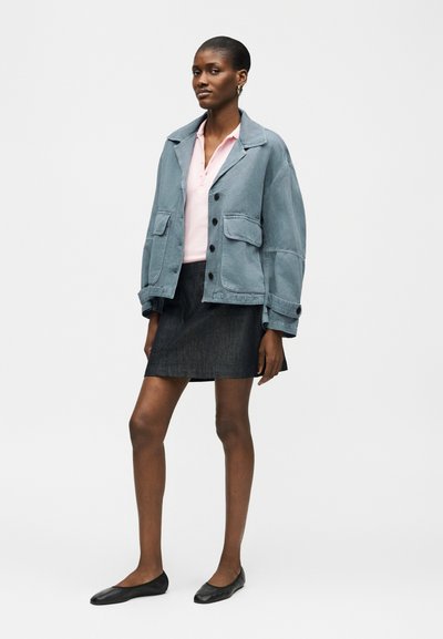 Femme portant une veste oversize bleu clair, une chemise rose pâle à col, une mini-jupe foncée et des ballerines noires, debout sur un fond blanc uni.