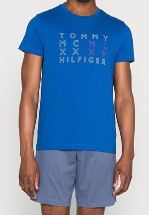 Homme portant un t-shirt bleu à manches courtes avec le texte "Tommy Hilfiger" et un short bleu foncé, debout devant un fond uni.