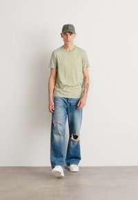 T-shirt à manches courtes vert clair, jeans bleus déchirés avec de grandes déchirures, baskets blanches et casquette verte. Design uni, coupe décontractée.