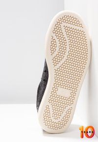 Semelle d'une chaussure noire avec semelle texturée couleur crème comportant des motifs de préhension circulaires, photographiée à la verticale sur un fond blanc en angle.