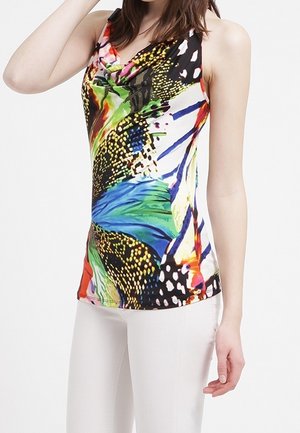 Femme vêtue d'un haut sans manches avec un motif abstrait coloré et un pantalon blanc, posant avec un bras levé et l'autre baissé.