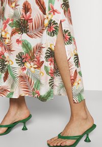 Gambe che indossano una gonna maxi floreale con uno spacco alto e sandali verdi con tacco e sottili cinturini su un pavimento chiaro.