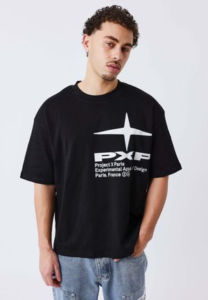 Uomo con capelli ricci che indossa una maglietta nera del Project X Paris e jeans azzurri, in piedi davanti a uno sfondo bianco uniforme.