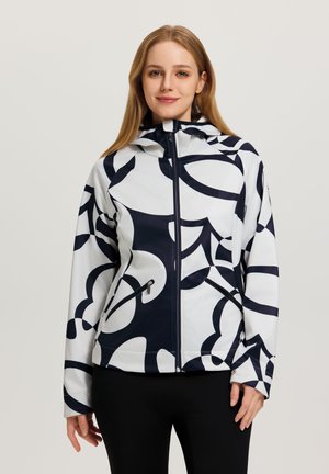 Frau trägt eine weiße und schwarze Jacke mit abstraktem Muster und Reißverschluss sowie Kapuze, steht vor einem schlichten hellen Hintergrund.