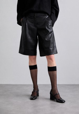 Personne portant un short en cuir noir jusqu'aux genoux, des bas noirs transparents montant jusqu'aux genoux et des chaussures Mary Jane noires sur un sol gris.