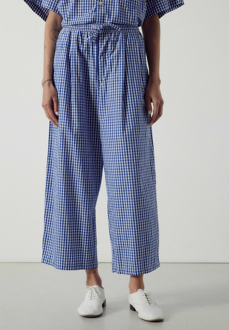 Femme portant un pantalon large à carreaux bleu et blanc, associé à des chaussures blanches à lacets, debout devant un fond uni.